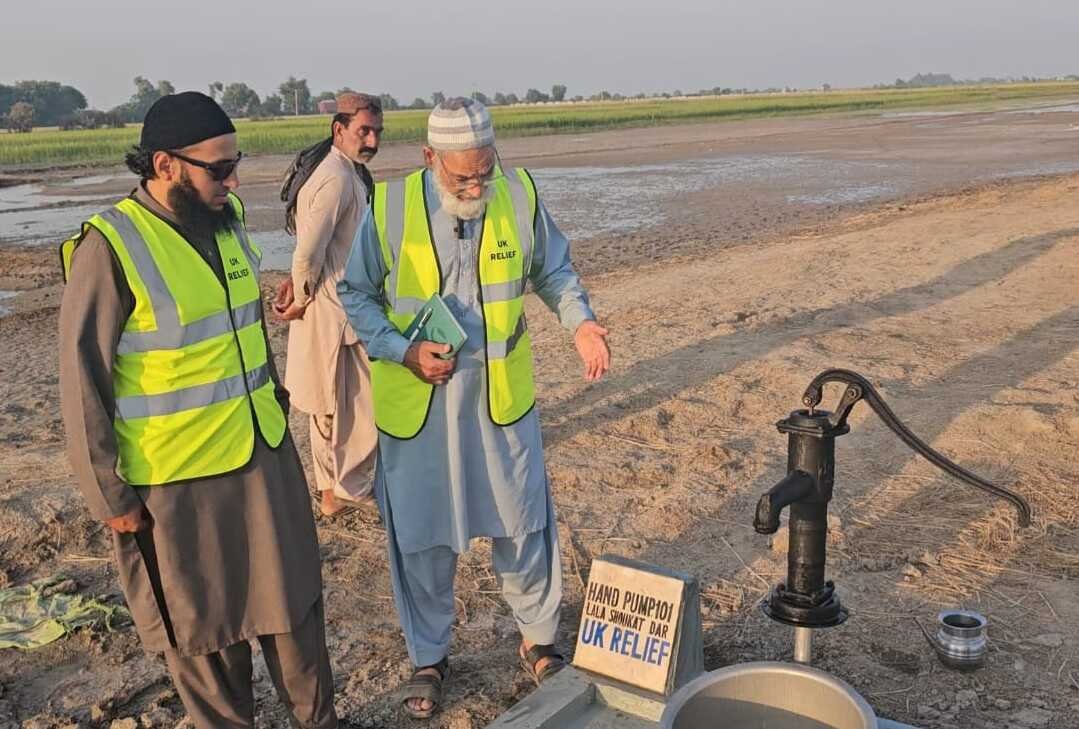 a group of men standing next to each other with hi vis jackets next to a water hand pump in a field